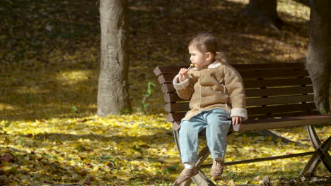 cute little girl sitting on a banch in autumn park enjoying sucking sweet lollipop candy outdoors - full body in slow motion