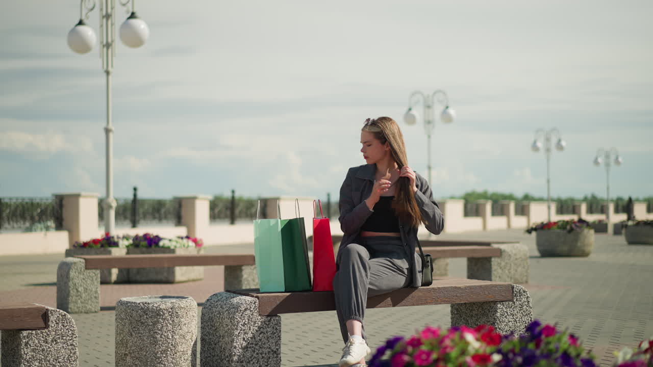Woman seated on bench with three colorful shopping bags beside her, crosses her legs while straightening her hair, she looks off into the distance in a outdoor setting with blooming flowers around