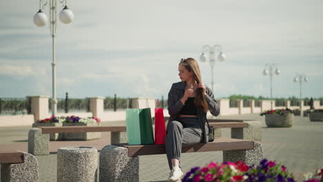 woman seated on bench with three colorful shopping bags beside her, crosses her legs while straightening her hair, she looks off into the distance in a outdoor setting with blooming flowers around