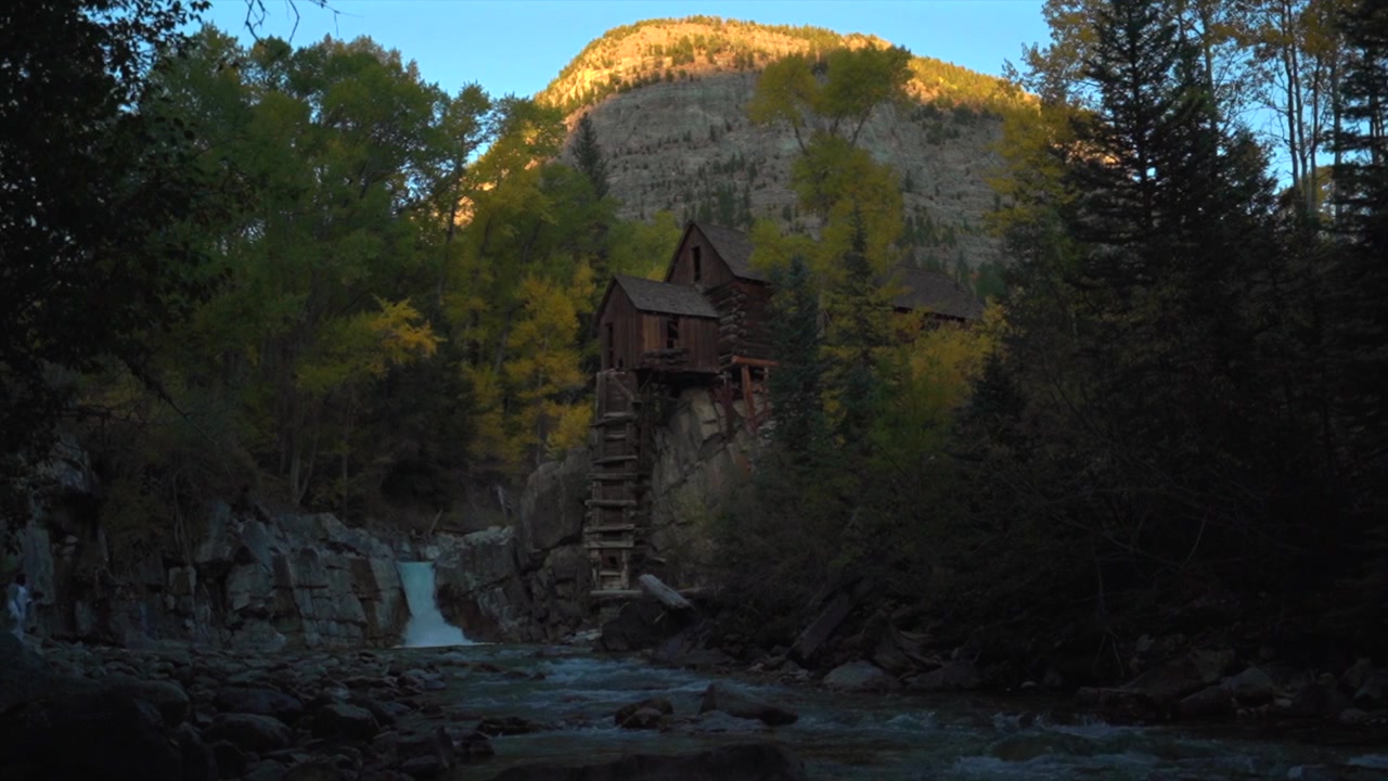 Il Crystal Mill, vecchio edificio in legno per l'estrazione dell'argento, centrale elettrica su affioramento roccioso, Marble, punto di riferimento del Colorado. Ripresa aerea con drone. Autunno, cascata del fiume Crystal, tramonto pomeridiano, cielo blu, paesaggio con movimento a sinistra.