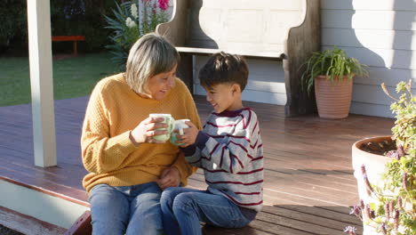 Happy-senior-biracial-grandmother-and-grandson-holding-mugs-sitting-on-terrace,-slow-motion