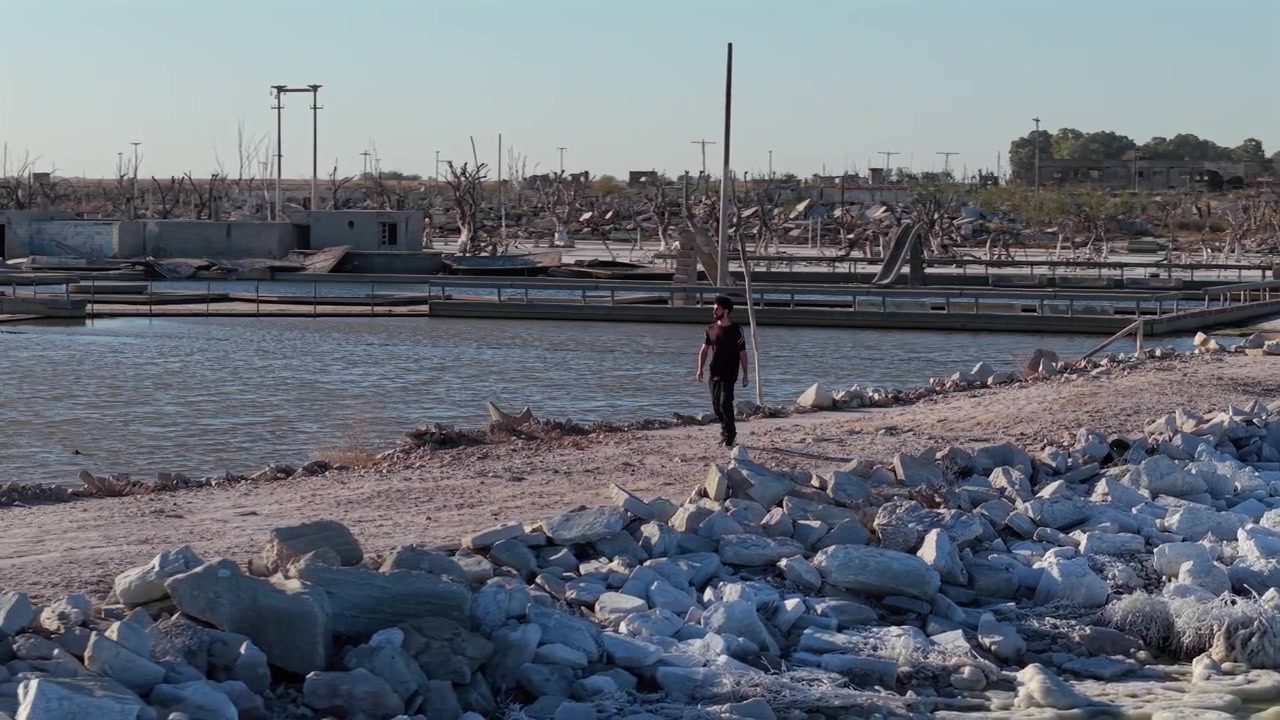 Arjantin, Buenos Aires'teki Villa Epecuén'in sular altındaki kalıntıları arasında yürüyen adam, yıkıcı selin yok ettiği eski tatil kasabasının tuzla kaplı kalıntılarının yanından geçiyor, ağır çekim drone takibi.