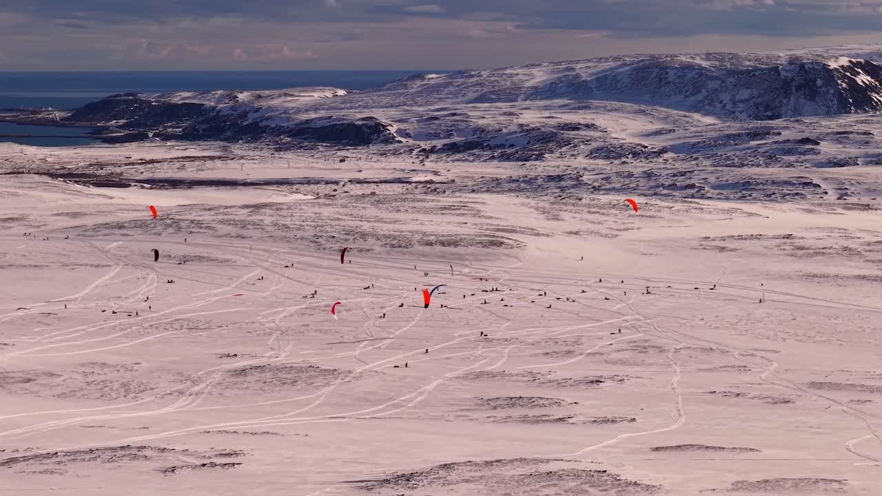 Aerial drone footage slowly flying around people who are kite surfing or skiining on winter white snow covered arctic Norway endurance race during a sunny evening. Barents sea visible with mountains.