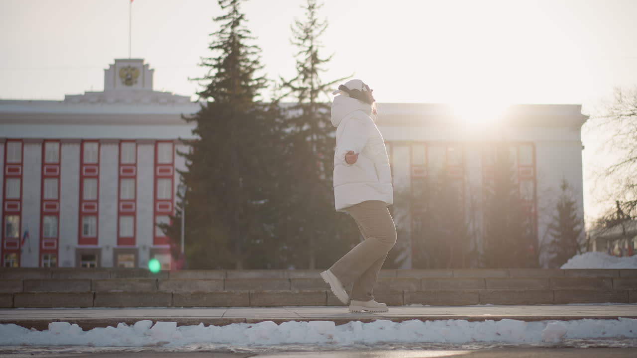 Girl dances gracefully on snow lined step pavement during winter sunset, immersed in choreography, wearing white coat and beanie, with sun flare casting warm glow behind government style building
