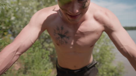 face view of tourist pushing up with full energy and strain on face, shirtless man training on grassy riverbank dirt slope, sand camo on cheeks, muscles flexed under bright daylight