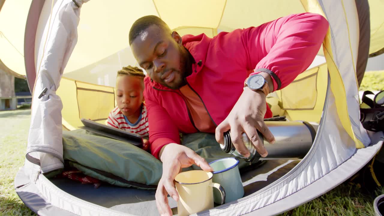 padre e hijo afroamericanos felices en la tienda usando tableta y vertiendo té en el jardín, en cámara lenta