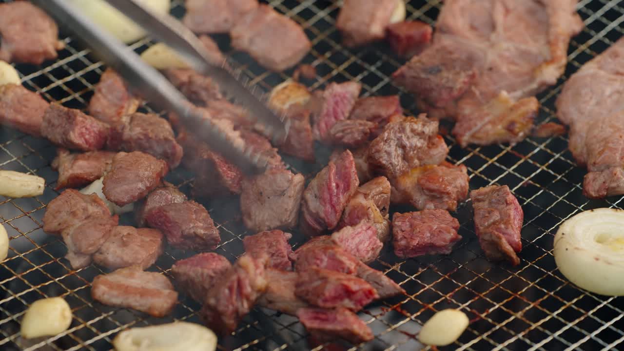 Extreme close-up, slow-motion shot of sizzling, juicy, bite-sized pieces of grilled beef and pork cooking with garlic over a hot charcoal fire during a Korean barbecue