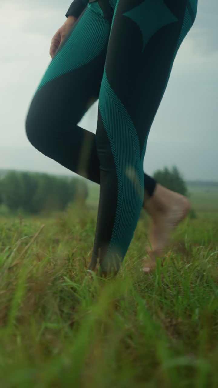 Leg view of a woman in black and green leggings walking barefoot on grassy field towards a mat on the ground, stepping onto the mat under a cloudy sky in a vast grassy field