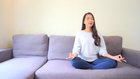 Junge-Brünette-Frau,-Die-Auf-Einem-Sofa-In-Ihrem-Wohnzimmer-Meditiert-Oder-Yoga-In-Lotuspose-Praktiziert