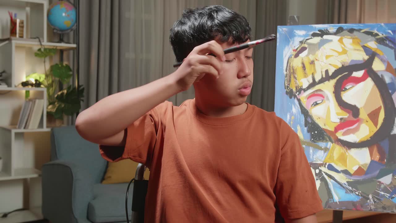 Asian Artist Boy In Wheelchair Holding Paintbrush Wipe The Sweat Before Crossing His Arms And Smiling While Painting A Girl On The Canvas