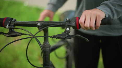 close-up of someone adjusting the handlebar of parked bicycle in grassy field, the person holds the handlebar with firm grip, checking the brake functionality while standing in the park