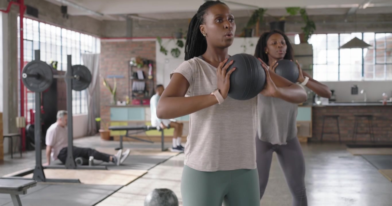 African American women following cue, holding black medicine balls, squatting on mats in loft gym
