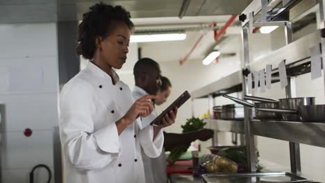 African-american-female-chef-taking-orders-and-using-tablet-in-restaurant-kitchen