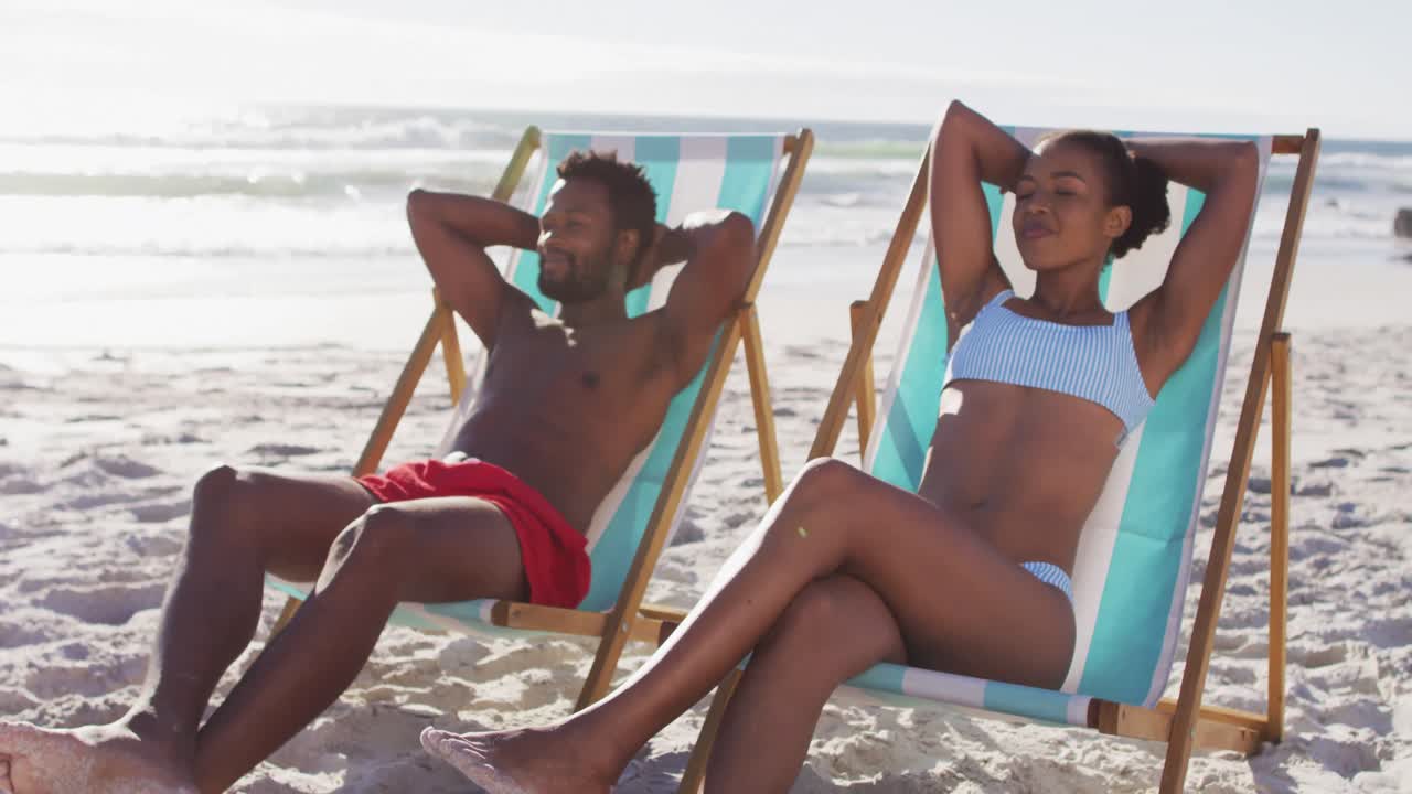African american couple sunbathing while sitting on deck chairs at the beach