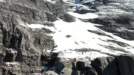 Drone-shot-flying-backwards,-revealing-a-young-man-sitting-down-on-the-edge-of-the-cliff-and-resting-next-to-a-big-wooden-cross-on-top-of-a-mountain-on-route-Via-Ferrata-in-Switzerland