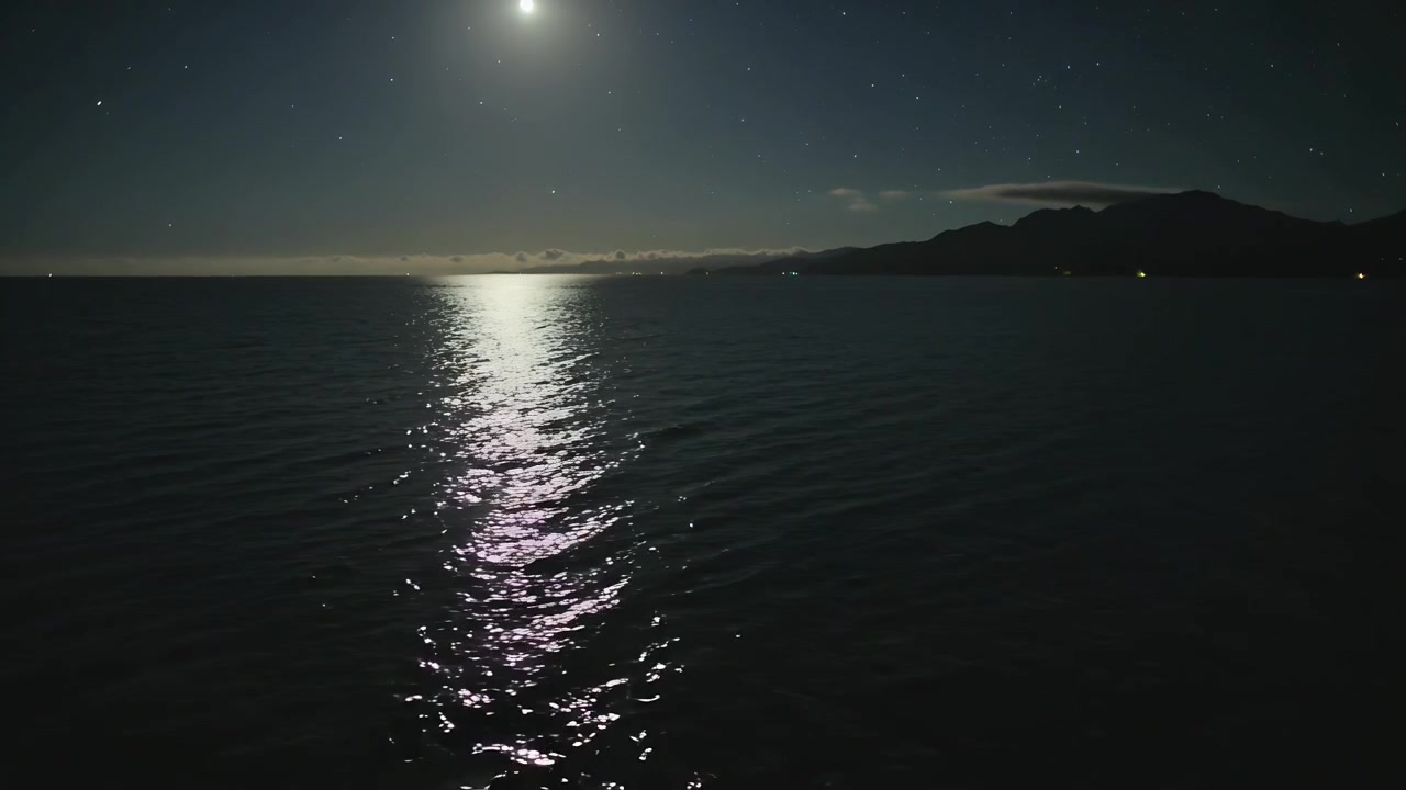 Riflesso tremolante della luna che si muove sull'acqua aperta di notte con leggere increspature e luci della riva.