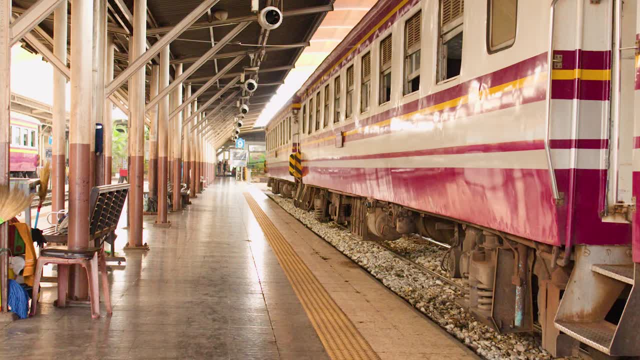 閑静な屋根付きのバンコク鉄道プラットフォームを走る、ピンク色のビンテージ旅客列車のトラッキングショット。