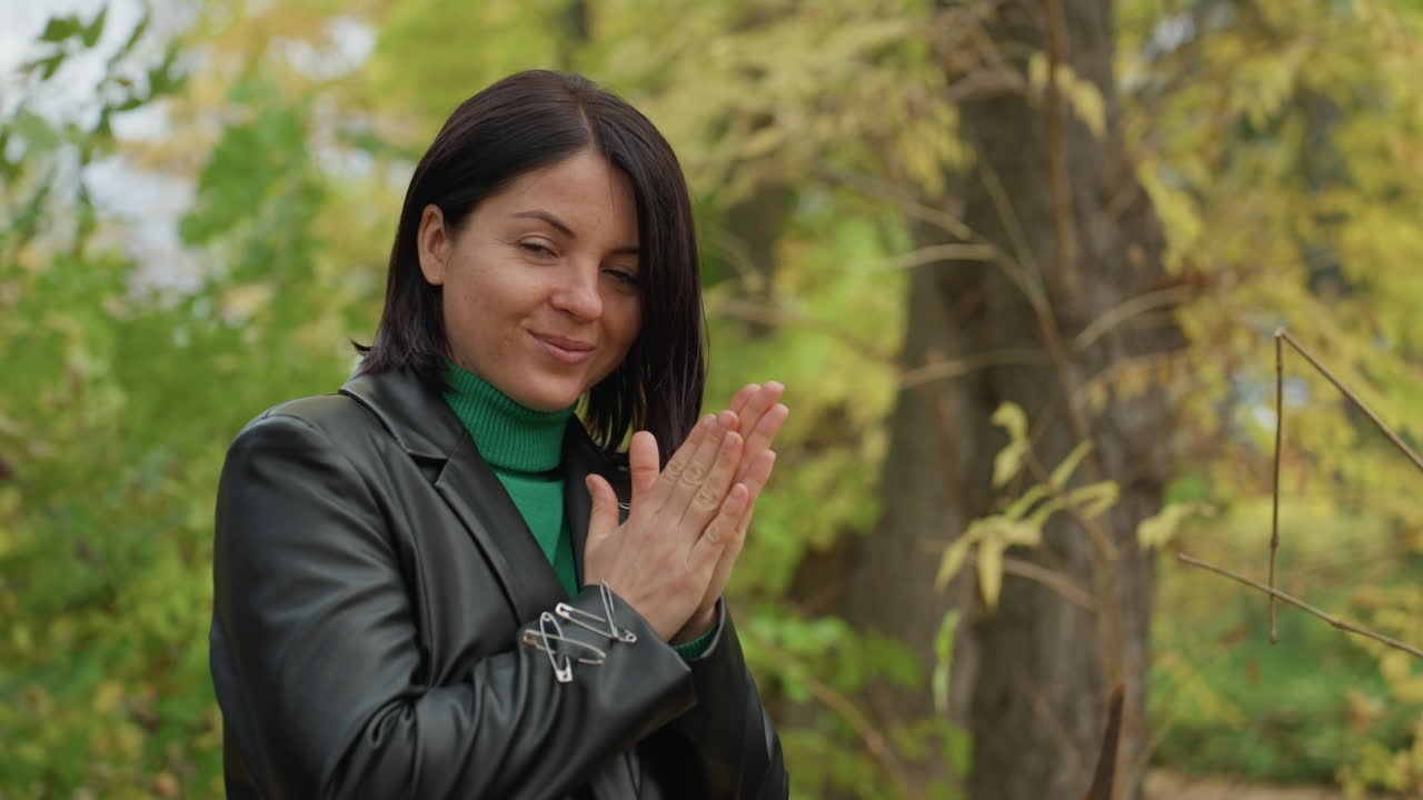 Mujer con chaqueta de cuero rezando en medio de un bosque otoñal con una bufanda verde y el pelo trenzado, manos entrelazadas, sonrisa sutil, ambiente contemplativo, apariencia de estrella de rock, serena soledad junto a los troncos de los árboles.