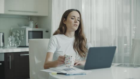 Mujer-Cansada-Usando-Computadora-Portátil-En-Casa.-Mujer-Seria-Bebiendo-Té-En-La-Cocina