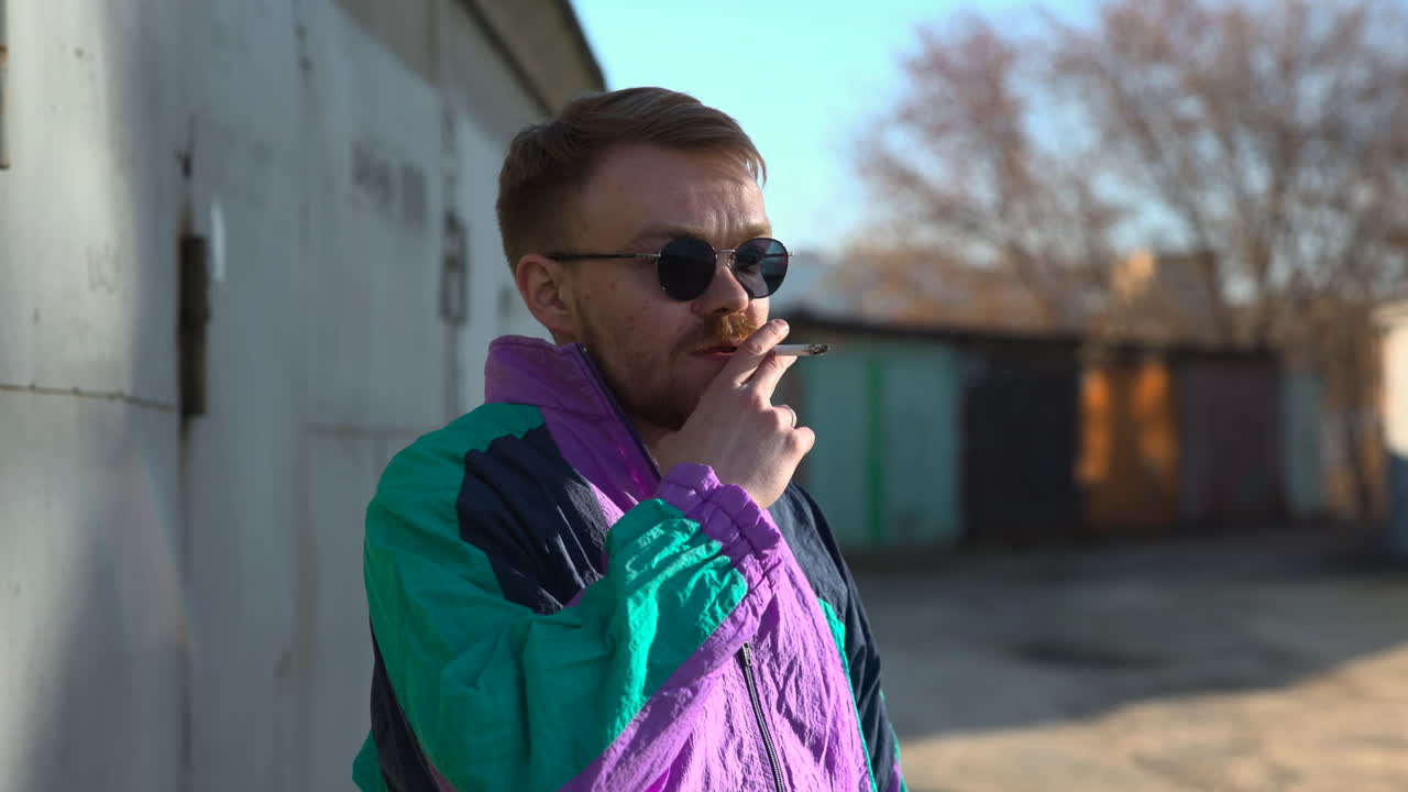 Hombre fumando pensativo cerca de puertas oxidadas, mano en los labios, mirada reflexiva, chaqueta vintage, pintura descascarada.