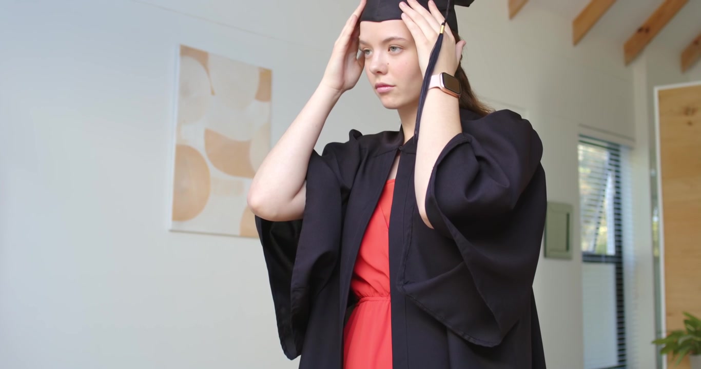Female adjusting red dress and gown, placing cap, smoothing tassel at home preparing for graduation