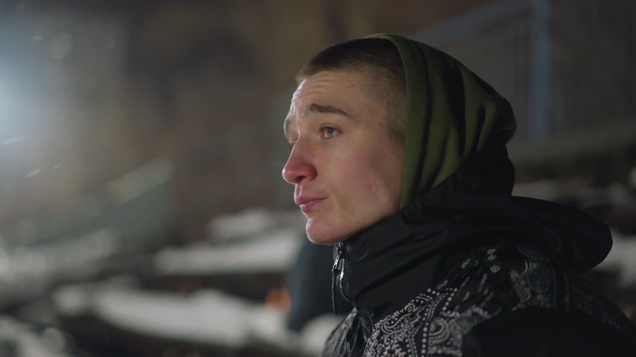 Close up of thoughtful teenage boy standing under bright light at night, looking around with serious expression as green lens flare highlights side of face against soft, blurred outdoor background