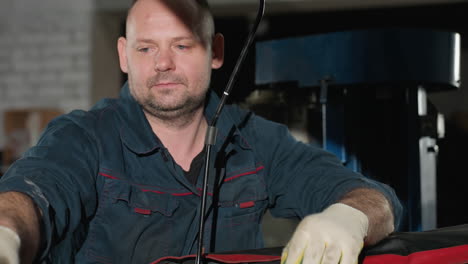 car repairer in blue uniform with gloves working on engine, exchanging tools with colleague in background, focused on repair task in a well-lit workshop environment with various tools and equipment