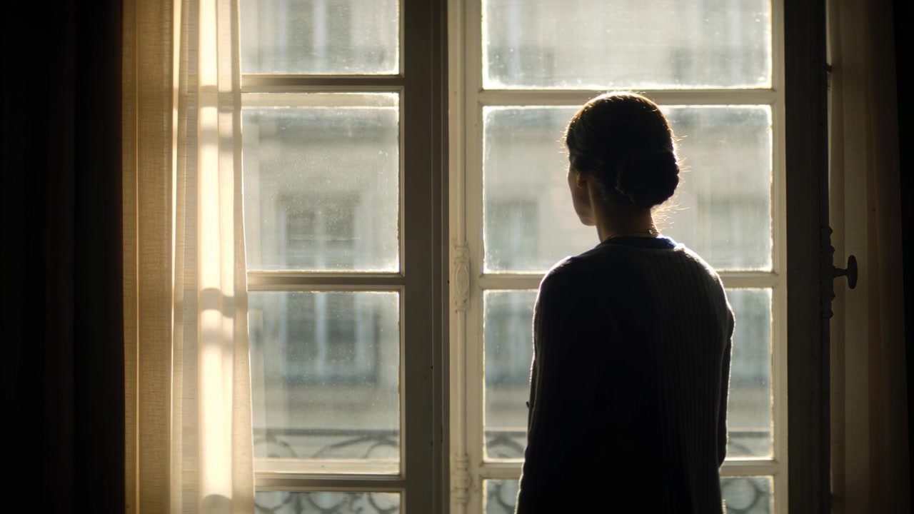 Sunlight drawing woman shifting toward window in flat, in dark sweater, sheer curtain, iron railing