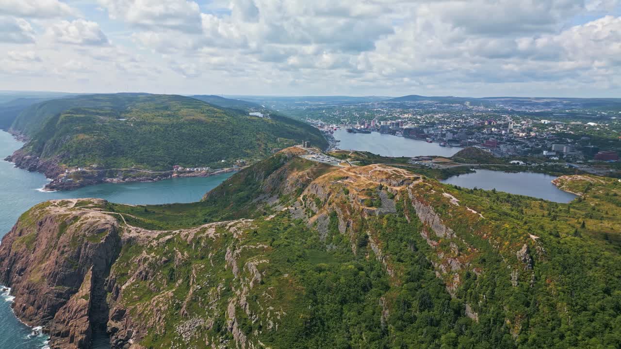 An aerial shot captures the undulating ridges and deep gullies of Signal Hill, with green slopes descending to a broad valley and distant harbour under a cloud-streaked sky