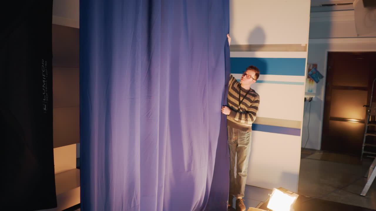 Male technician adjusting blue curtain in TV studio during set prep with floor lighting, striped backdrop and overhead rigging visible, cables trailing behind scenes broadcast production