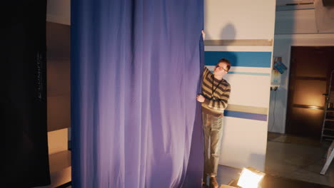 male technician adjusting blue curtain in tv studio during set prep with floor lighting, striped backdrop and overhead rigging visible, cables trailing behind scenes broadcast production