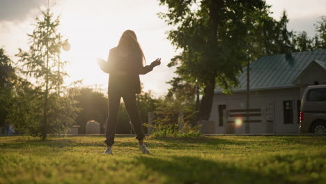 vista trasera de una dama bailando alegremente al aire libre en un campo cubierto de hierba con un libro en la mano, mientras la luz del sol crea una atmósfera radiante a su alrededor, la escena presenta vegetación, árboles y un edificio en el fondo