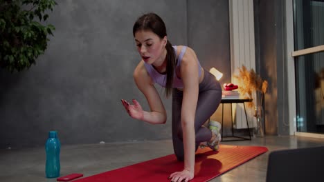 Confident-athletic-brunette-girl-in-a-purple-top-and-gray-pants-is-doing-sports-on-a-red-mat-using-video-lessons-using-laptops-in-a-modern-apartment-at-home