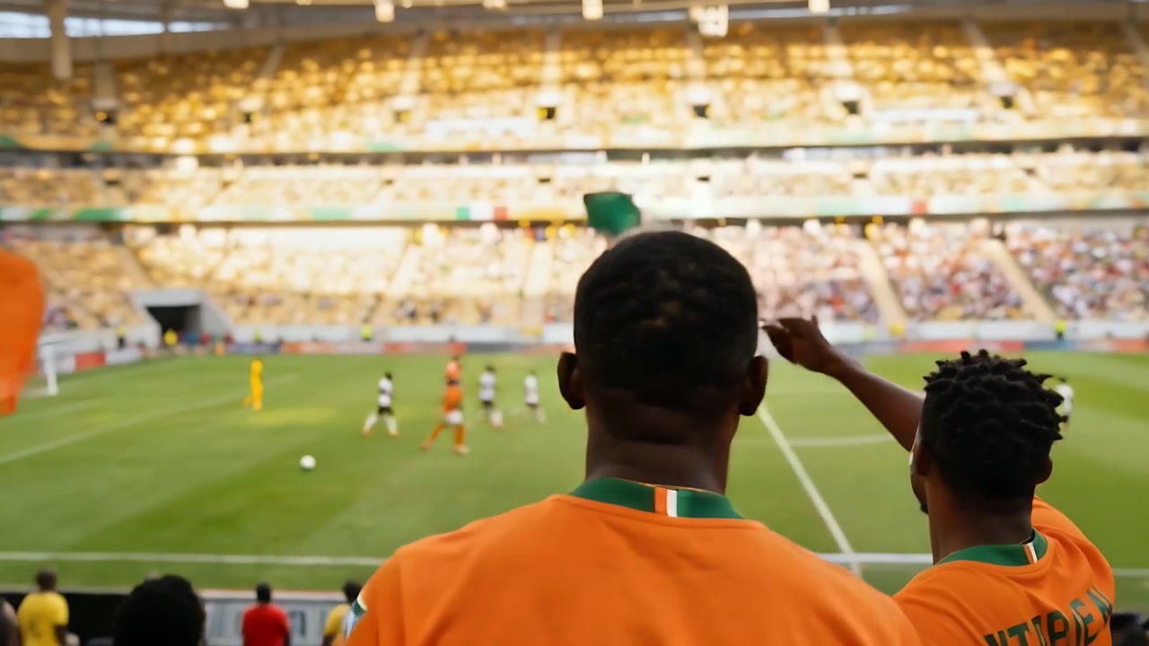 Tre tifosi con maglie arancioni sventolano alla ringhiera, tenendo bandiere tricolori per la squadra mentre il pallone si dirige verso la porta.