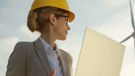 lower view of caucasian woman engineer wearing a helmet using laptop while checking operation of the windmill tourbines