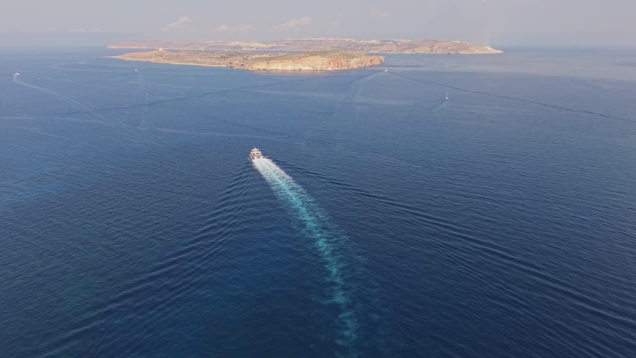 Drone footage of a motorboat cruises across the calm blue sea, leaving a long white trail behind. The distant coastline and cliffs create a serene horizon under a hazy sky