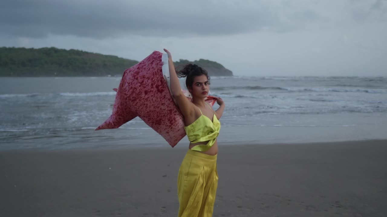 imágenes cinematográficas en cámara lenta de una modelo de moda india vestida con un traje amarillo en una playa de arena en un día ventoso sosteniendo una chaqueta de seda roja en el cielo, slomo, goa, india