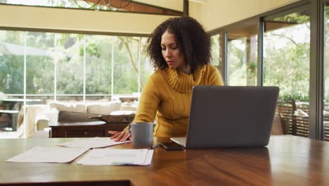 Mixed-race-woman-working-at-home-in-living-room,-using-laptop,-looking-at-paperwork