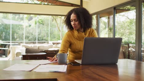 mujer de raza mixta trabajando en casa en la sala de estar, usando una computadora portátil, mirando papeles