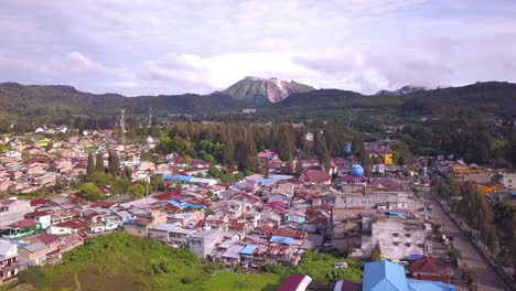 elevating drone shot from a balcony in berastagi town revealing mount sibayak surrounded by clouds, forest and pine trees in north sumatera, indonesia