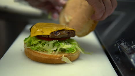 Trabajador-De-Comida-Rápida-Pone-Bollo-Encima-De-Hamburguesa-Con-Queso,-Cierre-Lento