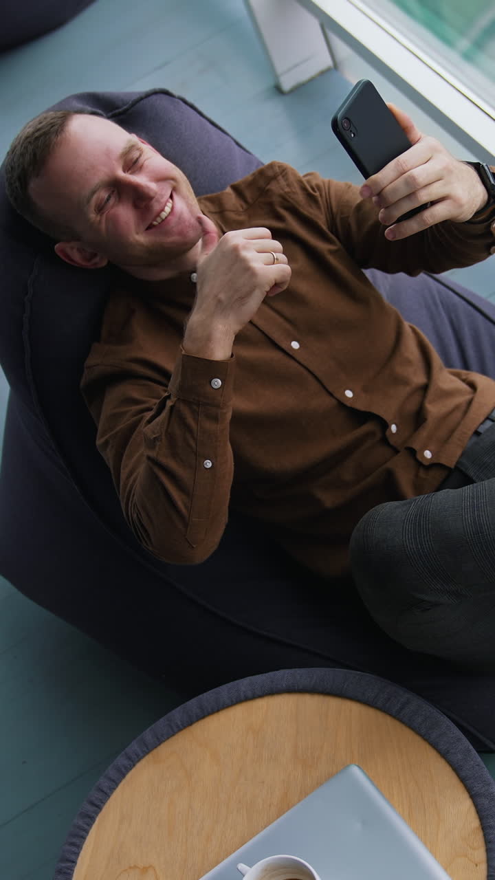 Businessman is having a video call. Young man in brown shirt and trousers sitting in a chair and talking online through the phone near the window. Top view. Vertical video