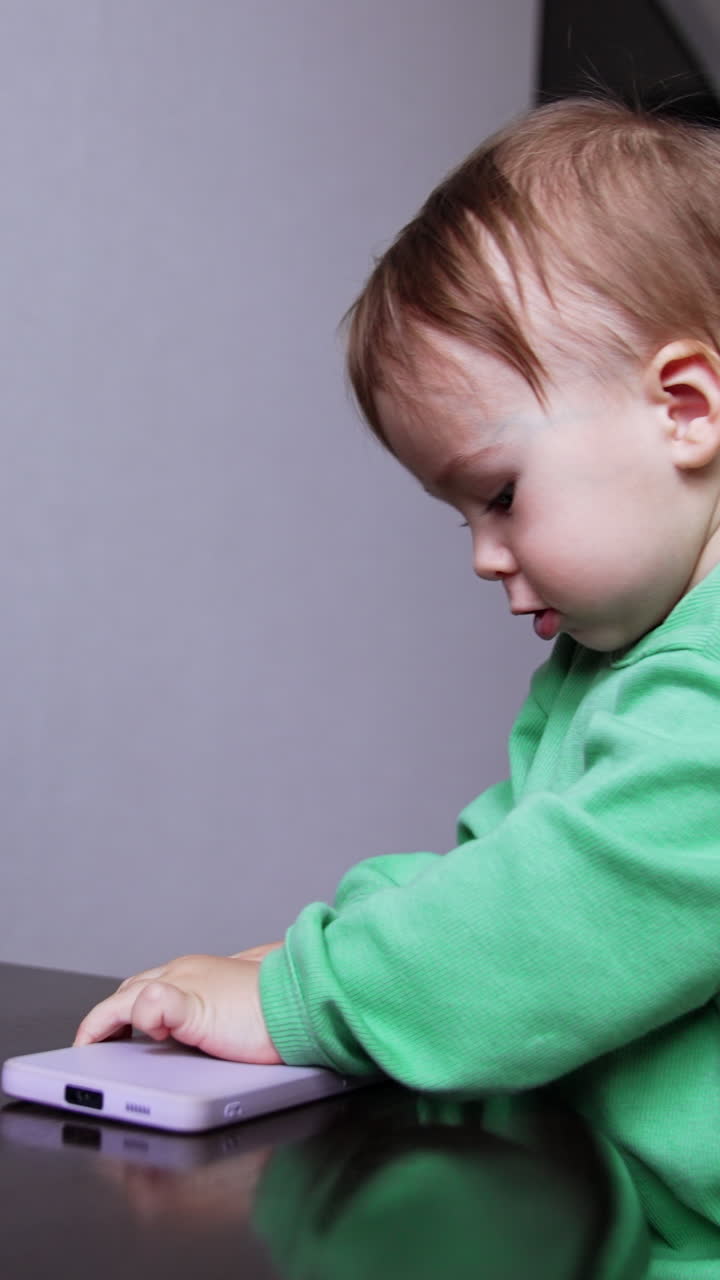 Lovely child of one year age playing with the phone on the stairs. Focused kid turning the gadget in hands curiously exploring it. Vertical video