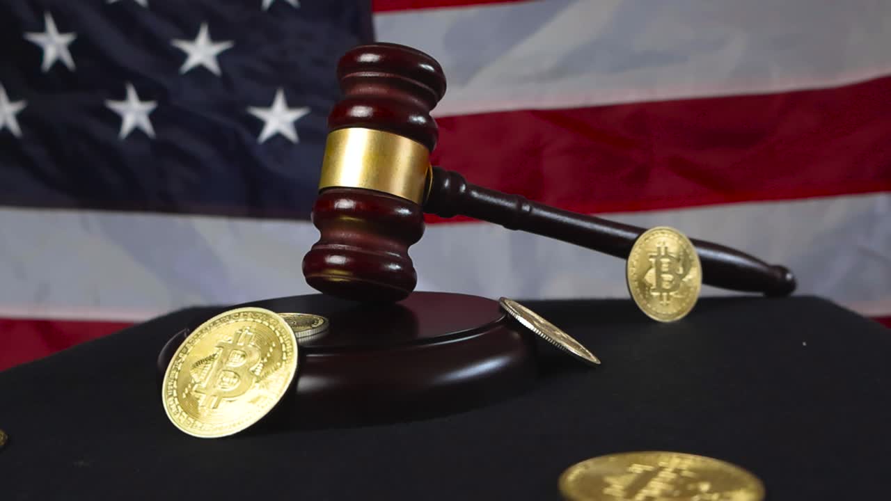 Brown wooden court hammer with bronze decorations placed next to golden shiny Bitcoin cryptocurrency money coins that are all in front of a United States of America flag while video orbits around them