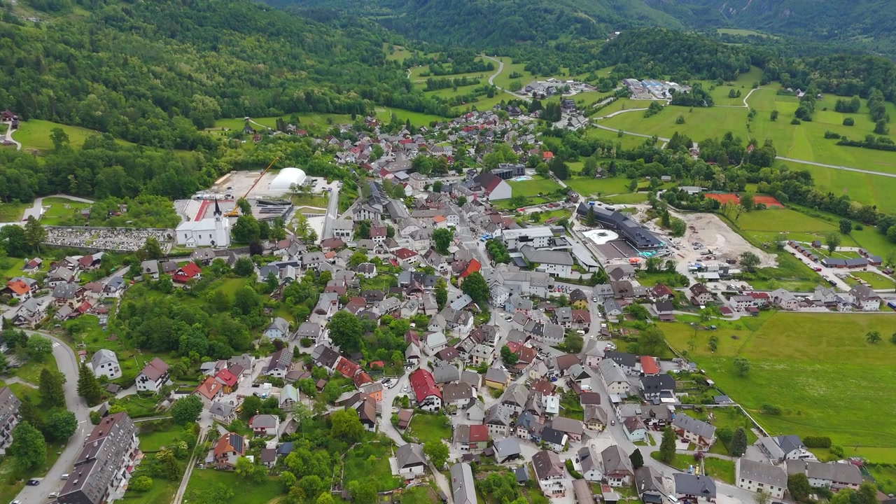 Alpebyen Bovec strækker sig ud over en grøn dal under De Juliske Alper i Slovenien. Huse med røde tage klynger sig langs snoede veje, omgivet af skovklædte bjergskråninger. Bredt dronebillede fra luften.