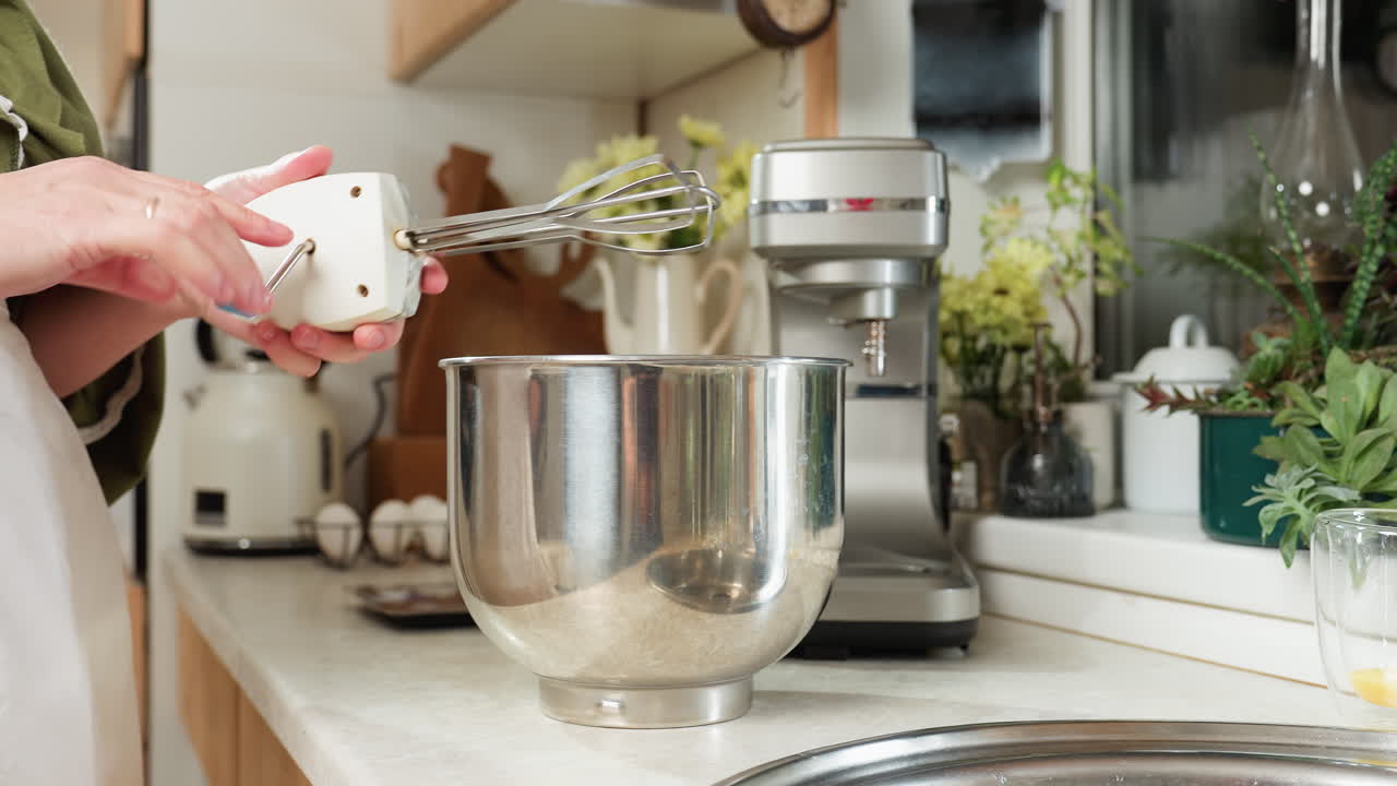 Close up of woman in green sleeve using handheld electric mixer inside stainless steel bowl in kitchen setting with mixer machine, plants, and glass with egg yolk on countertop