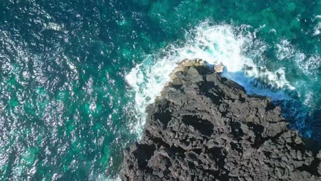 waves crashing on rugged black cliffs surrounded by turquoise ocean waters , aerial view