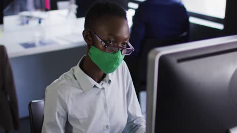 African-american-woman-wearing-face-mask-using-computer-while-sitting-on-her-desk-at-modern-office