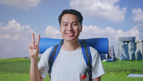 close up of asian male hiker with mountaineering backpack smiling and showing peace gesture while traveling in stonehenge
