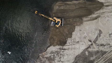 drone rises to reveal an excavator removing sediment from a river flowing on sandy bank